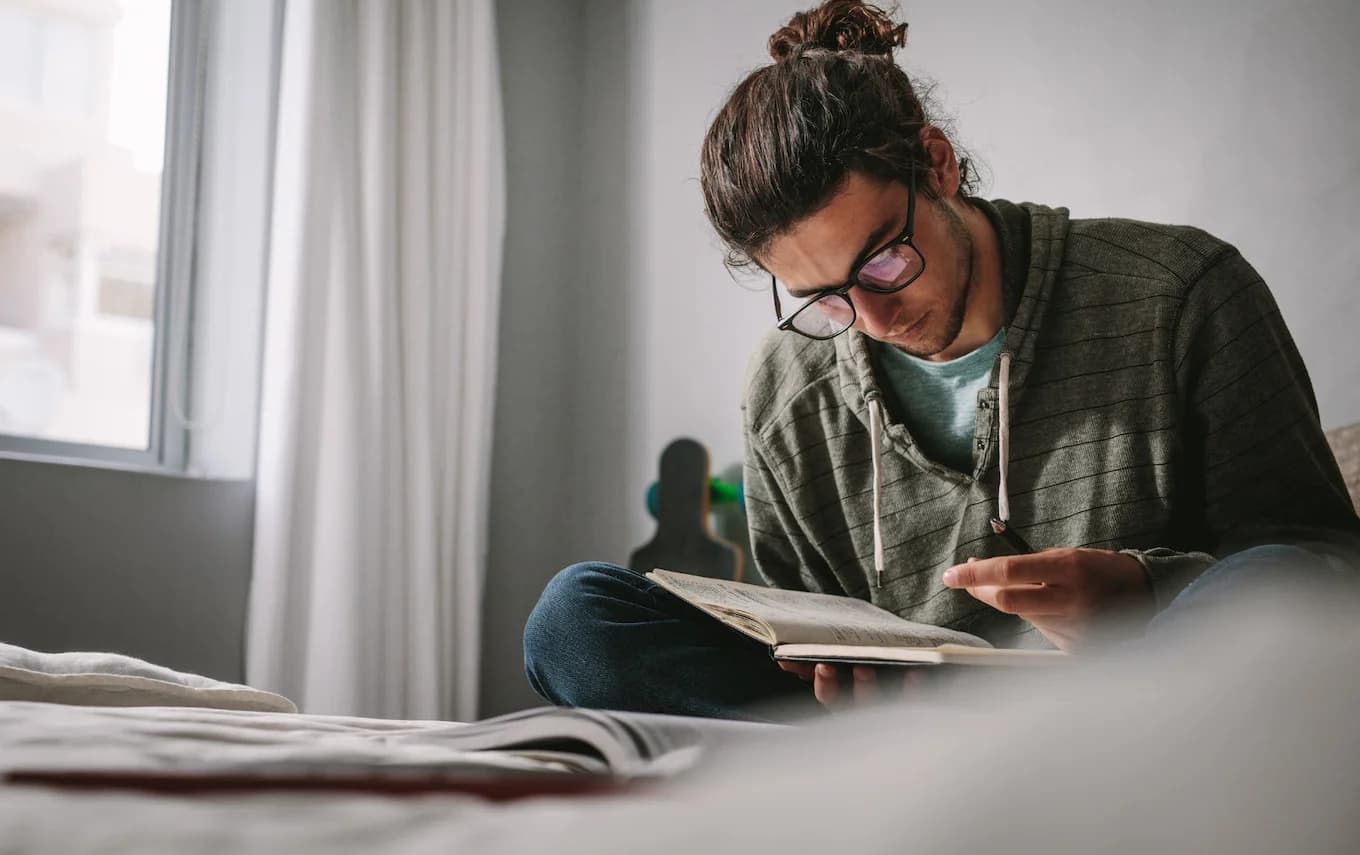 Giovane studente con occhiali concentrato nella lettura di un libro, seduto vicino a una finestra illuminata dalla luce naturale.