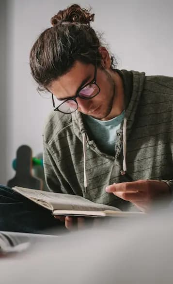 Giovane studente con occhiali concentrato nella lettura di un libro, seduto vicino a una finestra illuminata dalla luce naturale.