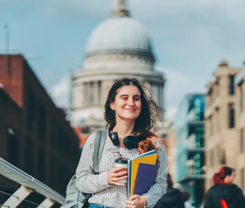 Giovane studentessa sorridente con cuffie e libri in mano, cammina per la città con uno sfondo urbano suggestivo.